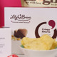 Close up. A box of Lily O'Brien's Crispy Hearts chocolates is prominently displayed. In front, there is a bowl filled with yellow snack chips. To the left, a white box labeled "Candle" is partially visible. The background features a pink surface, adding color to the composition. Close up. A box of Lily O'Brien's Crispy Hearts chocolates is prominently displayed. In front, there is a bowl filled with yellow snack chips. To the left, a white box labeled "Candle" is partially visible. The background features a pink surface, adding color to the composition.